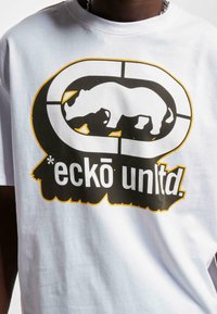 Bijela pamučna majica s velikim crnim i žutim grafičkim prikazom siluete nosoroga unutar kružnog dizajna te tekstom „eckō unlt.d.“