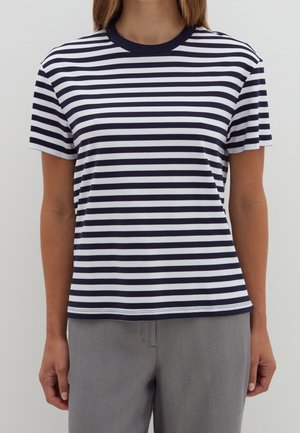 Person iført en marineblå og hvid T-shirt med korte ærmer og vandrette striber og grå bukser, stående mod en ensfarvet baggrund.