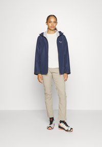 Marine vanntett jakke med grå hette, kombinert med beige bukser og joggesko med svarte, hvite og oransje detaljer.
