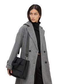 Borsa a spalla nera trapuntata con patta a ribalta, accessori in argento e tracolla regolabile, tenuta da una persona che indossa un cappotto grigio.