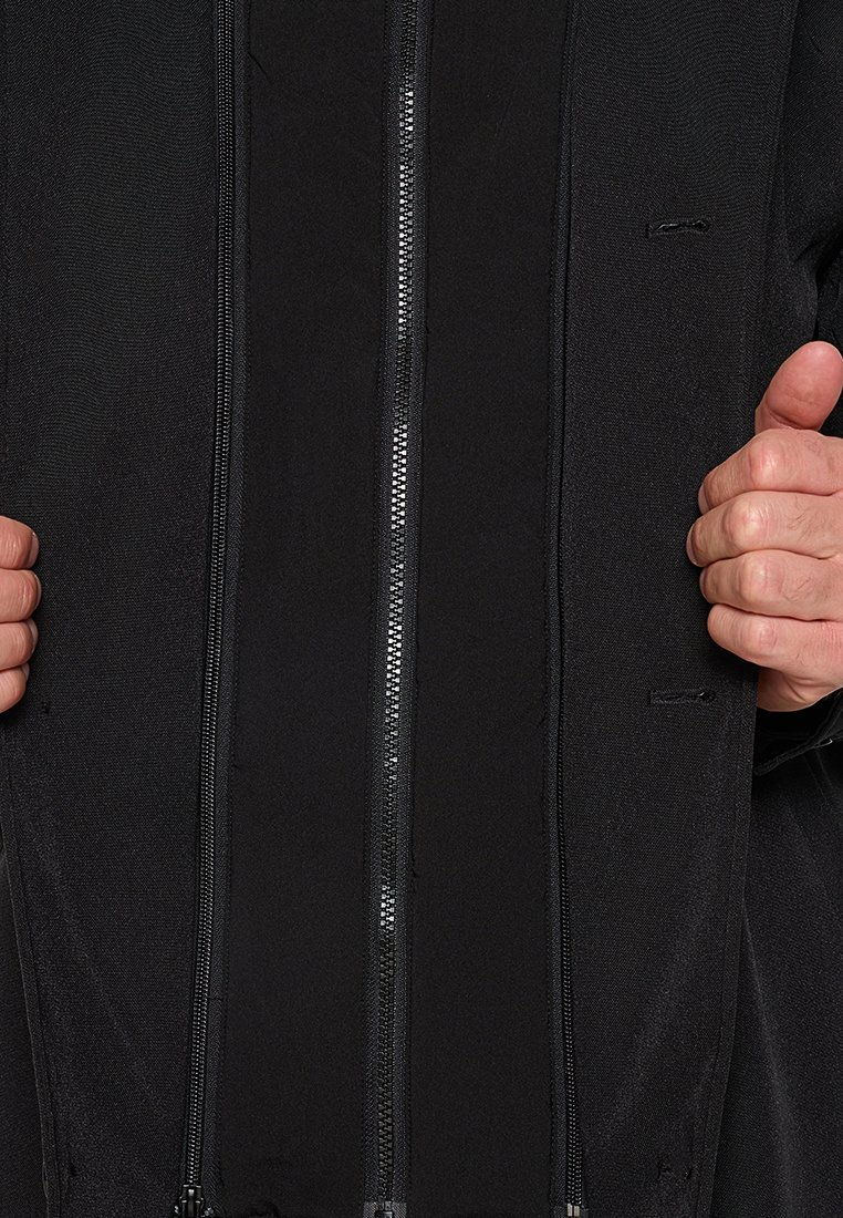 Zwarte jack met een centrale rits, gestructureerde stof en dubbele zijzakken. Het ontwerp is minimalistisch zonder zichtbare patronen of logo's.
