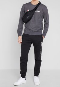Sudadera gris de cuello redondo con el logo blanco de "Champion", pantalones de chándal negros con logo lateral, zapatillas blancas y un bolso de bandolera negro.