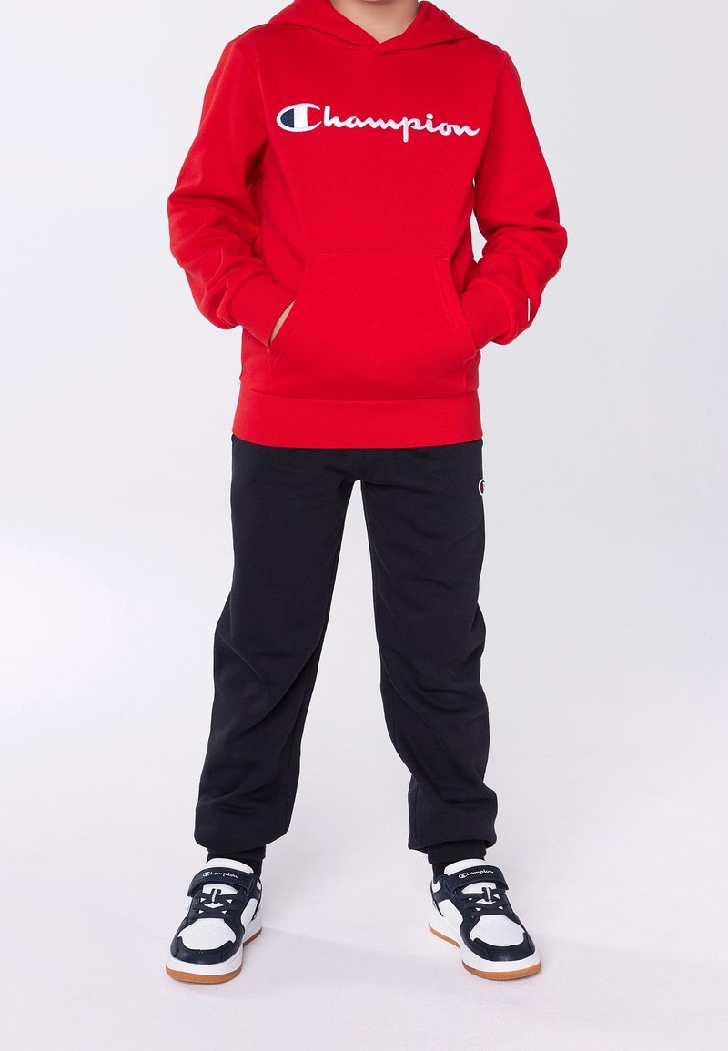 Sudadera roja con capucha y el logo blanco de "Champion", bolsillo frontal, combinada con pantalones de chándal negros y zapatillas en negro y blanco con suela de goma.