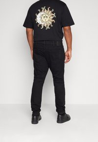 Camiseta negra con un gráfico de sol y luna en la espalda, combinada con jeans negros y zapatos negros, mostrando un conjunto casual desde atrás.