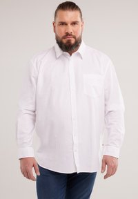 Man with beard and tied-back hair wearing a white long-sleeve button-up shirt and blue jeans, standing against a plain background.