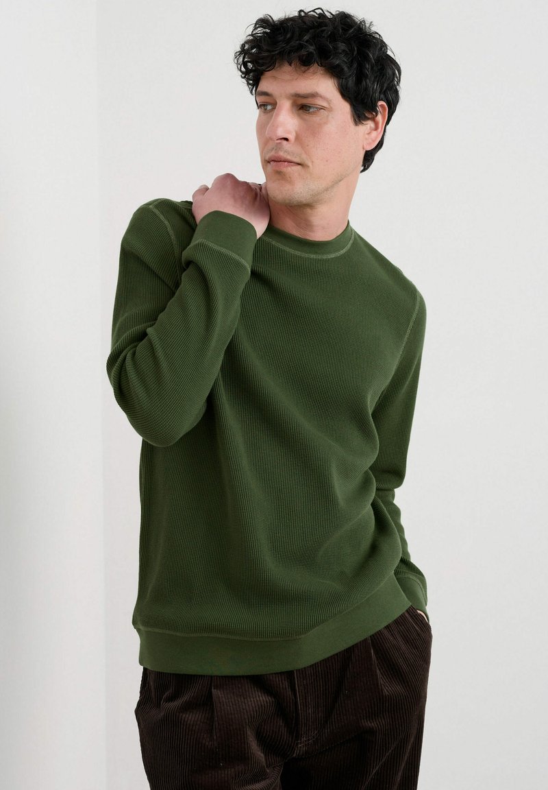 Grüner gerippter Pullover mit langen Ärmeln, rundem Ausschnitt und strukturierter Oberfläche. Kombiniert mit braunen Cordhosen.
