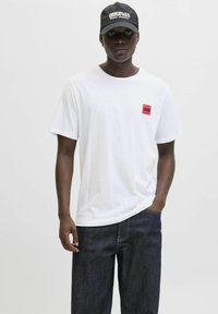 Hvid bomulds t-shirt med korte ærmer og en rød mærket firkant på venstre bryst, parret med mørk denim jeans og en sort kasket.