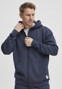 Marine blauwe zip-up hoodie met een trekkoord capuchon, kangoeroezakken en een klein logo op de linkerkant van de borst. Gemaakt van zachte stof met een gladde textuur.