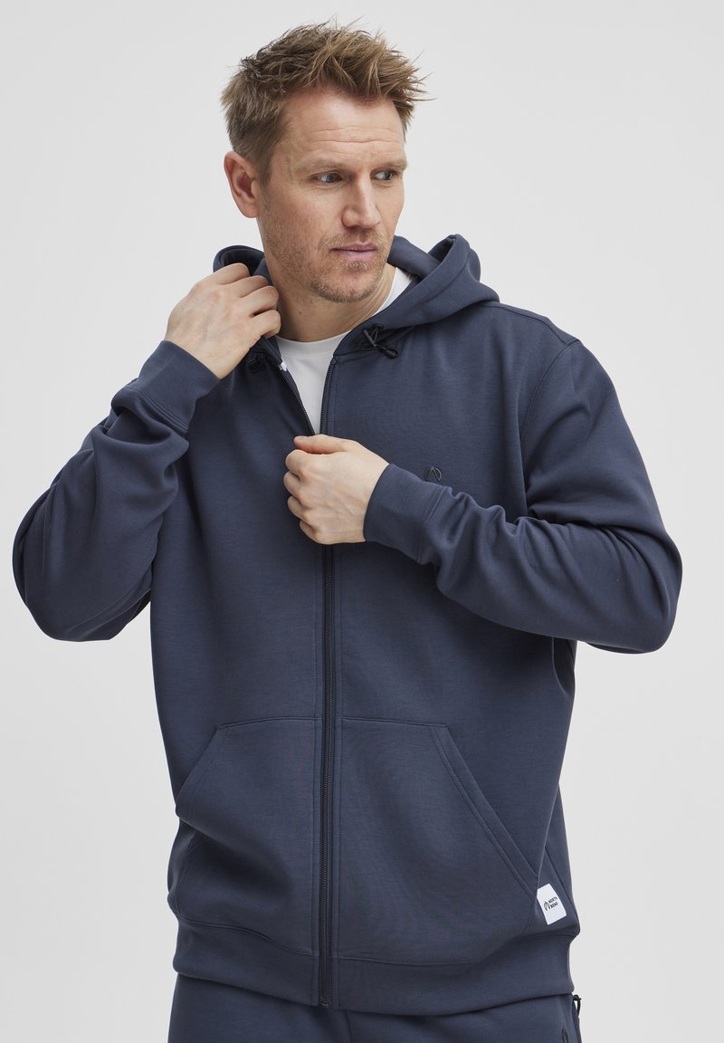 Marine blauwe zip-up hoodie met een trekkoord capuchon, kangoeroezakken en een klein logo op de linkerkant van de borst. Gemaakt van zachte stof met een gladde textuur.