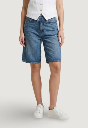 Persona che indossa pantaloncini di jeans blu di media lunghezza, gilet bianco con bottoni e sneakers bianche, in piedi davanti a uno sfondo chiaro e semplice.