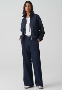 Marineblauwe cropped jacket met twee voorzakken, gecombineerd met donkere denim broeken met wijde pijpen en grijze sneakers, tegen een neutrale achtergrond.