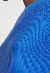 Niebieski sweter dzianinowy o fakturowanym, prążkowanym wzorze. Dekolt jest okrągły, a materiał wygląda na miękki i przewiewny.