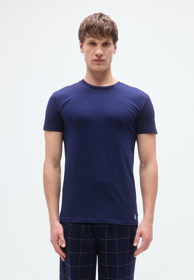 Μπλε ναυτικό κοντομάνικο μπλουζάκι από μαλακό ύφασμα με στρογγυλή λαιμόκοψη και μικρή λεπτομέρεια με λογότυπο στο τελείωμα, φορεμένο με καρό παντελόνι πιτζάμας.