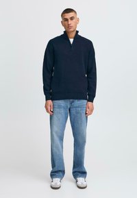Maglione blu navy con zip al collo, texture a maglia, abbinato a jeans dritti azzurri e sneakers bianche. Look semplice e casual.