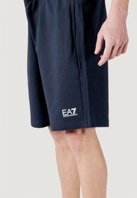 Shorts en coton bleu marine à coupe décontractée, avec poches latérales et un logo blanc "EA7 EMPORIO ARMANI" sur la partie inférieure de la jambe.