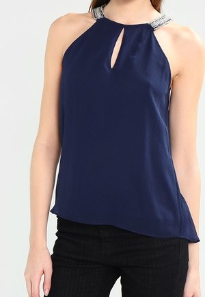 Vrouw draagt een mouwloze marineblauwe haltertop met zilverkleurige kralenbandjes en een keyhole-uitsnijding aan de voorkant, gecombineerd met een zwarte broek.