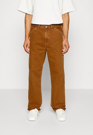 Homme portant un pantalon marron ample avec des poches avant et des baskets blanches, debout devant un fond blanc uni.