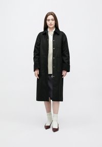 Femeie purtând o haină lungă neagră peste un pulover bej și o cămașă albă, asortată cu o fustă mov, șosete albe și pantofi burgunzi cu vârf ascuțit.