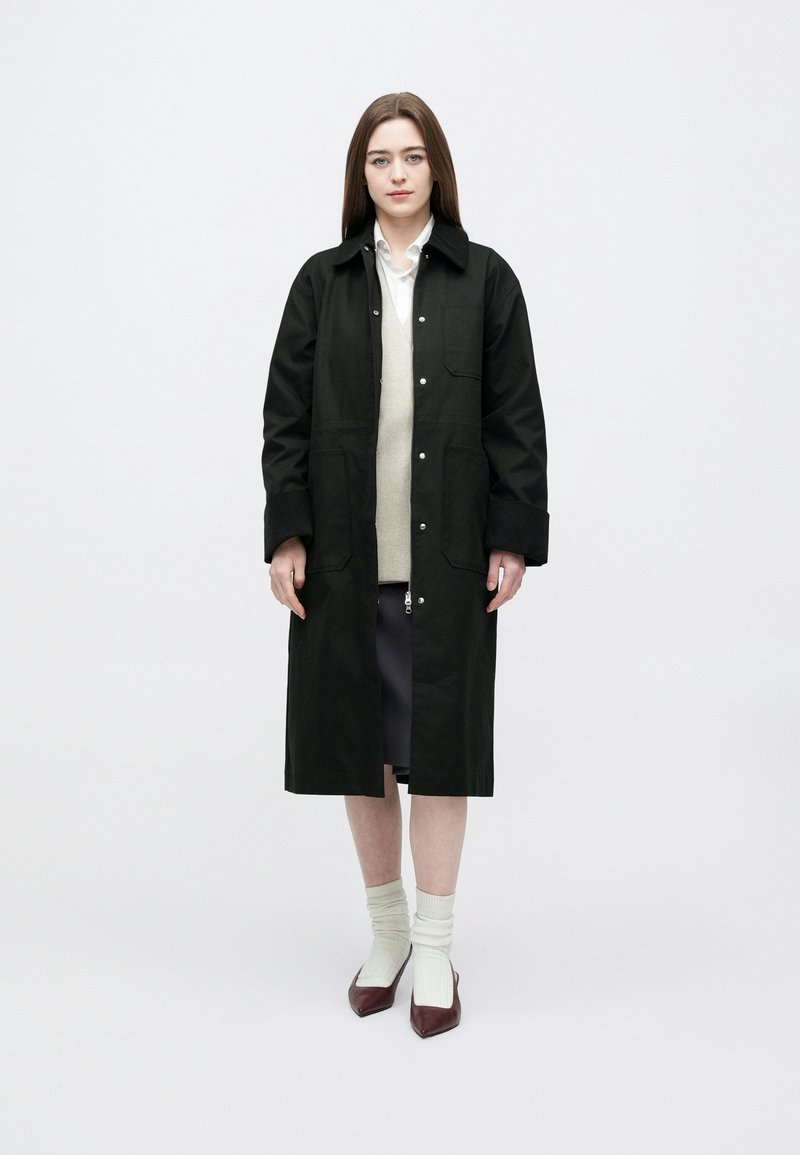 Femeie purtând o haină lungă neagră peste un pulover bej și o cămașă albă, asortată cu o fustă mov, șosete albe și pantofi burgunzi cu vârf ascuțit.