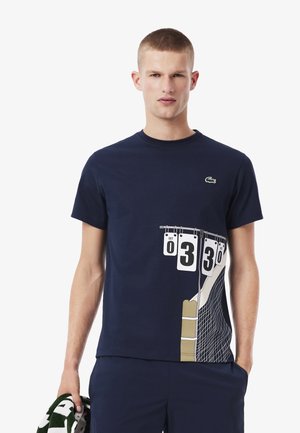 Giovanotto con una maglietta blu navy con una grafica di un tabellone di tennis e rete, che tiene un asciugamano verde e bianco nella mano sinistra.