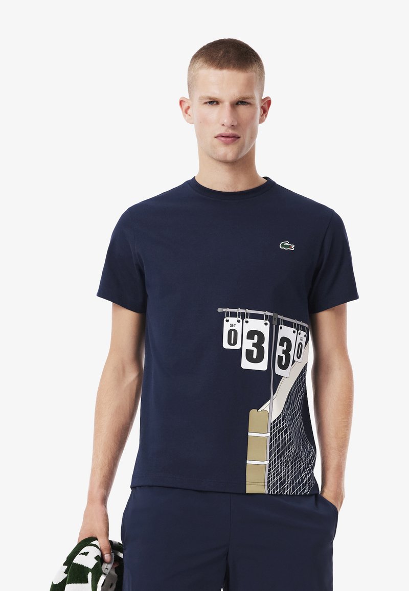 Joven vistiendo una camiseta de color azul marino con un gráfico de marcador de tenis y red, sosteniendo una toalla verde y blanca en su mano izquierda.