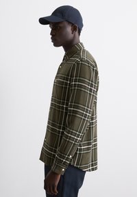 Camicia a quadri verde oliva con maniche lunghe, colletto abbottonato e dettagli in oro. Indossata sotto un cappellino da baseball blu navy.