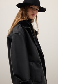 Black leather jacket with a shearling collar, large front pocket, and relaxed fit. The surface is smooth with a matte finish.