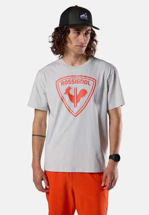 Uomo che indossa una maglietta grigia Rossignol con logo rosso, pantaloni arancioni, cappellino nero e smartwatch nero, in piedi con espressione neutra.