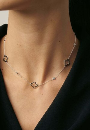 Silberne Halskette mit floralen Formen und durchbrochenen Designs sowie einem zentralen runden Akzent. Wird auf einem freien Hals über einem dunklen Oberteil getragen.