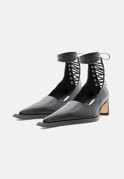 Μαύρα μποτάκια με μυτερή μύτη, χαμηλά χοντρά τακούνια και δερμάτινα κορδόνια που δένονται γύρω από τους αστραγάλους σε λευκό φόντο.