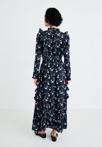Vestido longo preto com padrão floral colorido, mangas com volantes, cintura franzida e bainha em camadas com volantes; combinado com sapatos pretos.