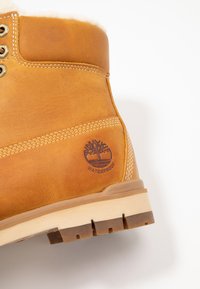 Wasserdichte, sandfarbene Lederstiefel mit hohem Knöchel, beigefarbenem Fellfutter, weißer Ziernaht und einem Baum-Logo, das an der Seite geprägt ist. Gummisohle.