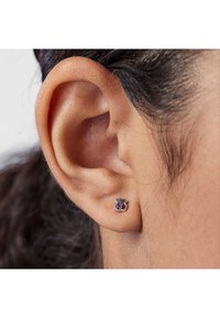 Primo piano di un orecchio di una persona con capelli scuri che indossa un piccolo orecchino a forma di orsacchiotto in argento con gemme viola.