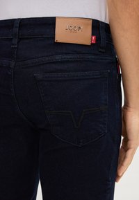 Jeans blu scuro con un'etichetta in pelle con la scritta "JOOP! JEANS." Presentano una cucitura a V sulla tasca posteriore e un design dal taglio classico.