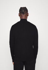 Suéter negro de canalé con cuello alto, mangas largas y un corte holgado. El diseño presenta una parte trasera sin costuras y un sutil detalle texturizado.