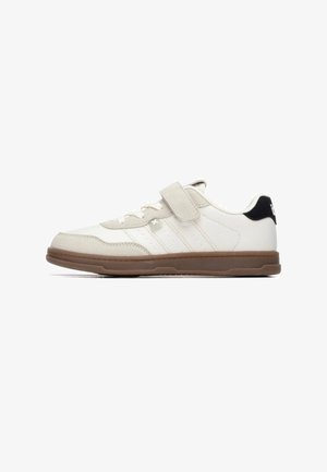 Zapatilla blanca y beige con puntera de ante, ventilaciones de malla, tres franjas y suela de goma marrón. Correa ajustable y detalle negro en el talón.