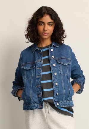 Tânără cu păr creț purtând o jachetă albastră din denim peste o cămașă cu dungi gri și albastre și pantaloni gri deschis, cu mâinile în buzunare.