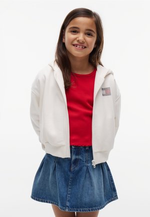 Fille souriante aux longs cheveux bruns portant un sweat à capuche blanc zippé, un haut rouge et une jupe en jean bleue, debout devant un fond blanc uni.