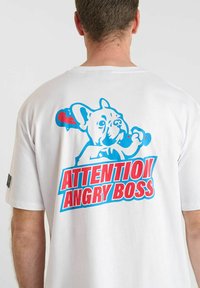 Man in een wit T-shirt met een blauwe en rode graphic van een hond die een knuppel vasthoudt, met de tekst "Let op, Boze Baas" op de rug.