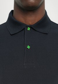 Gros plan d'une personne à la peau foncée portant un polo noir avec des boutons en forme de trèfle verts au niveau du col et de la poitrine.