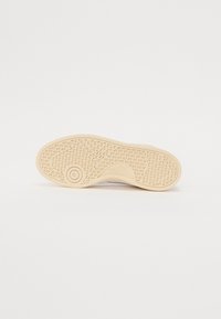 Suela de zapatilla en goma beige con un patrón de agarre texturizado, que presenta un acento circular y un logotipo. Diseño limpio y minimalista para una mejor tracción.
