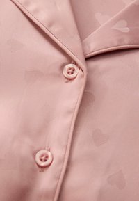 Haut de pyjama rose clair en tissu lisse, avec un motif de cœurs subtil, un col en V et deux boutons à l'avant.