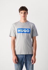 Pilkas medvilninis marškinėliai trumpomis rankovėmis, su ryškiai mėlyna stačiakampėmis dėžutėmis, kurioje balta spalva parašyta "HUGO." Klasikinio kirpimo dizainas.