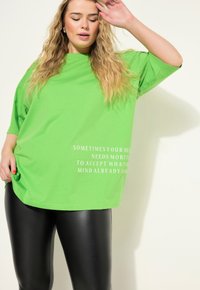Camiseta de gran tamaño de color verde brillante con escote redondo, con texto blanco en la parte frontal. Combinada con leggings negros y brillantes.