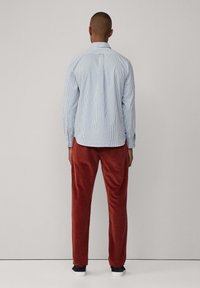 Camisa a rayas azul y blanca con dobladillo curvado, llevada con pantalones de pana color óxido, vista desde la parte trasera, combinada con zapatos oscuros.