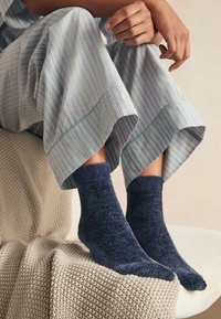 Mørkeblå bløde sokker med en blød struktur, båret med grå pyjamasbukser med striber og grønne accenter, hvilende på et struktureret tæppe.