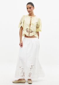 Rumena skrajšana bluzica z čipkastimi detajli, zavezana spredaj. Bela maksi krila z zapletenimi izrezi na robu. Bež ravne čevlje.