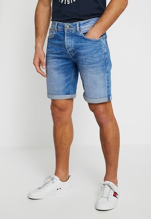 Homme portant un short en jean bleu retroussé, un t-shirt noir et des baskets blanches, debout avec une main dans la poche.