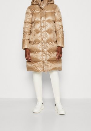 Lesklý opálený prošívaný kabát délky po kolena s kapucí, předním zipem a bočními kapsami, nošený s bílými legínami a bílými teniskami.