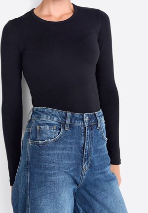 Frau trägt ein eng anliegendes schwarzes Langarmshirt, das in hoch taillierte blaue Jeans aus Denim mit Fronttaschen gesteckt ist.